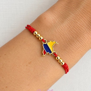 May include: A red cord bracelet featuring a central gold-toned metal charm shaped like the map of Colombia, filled with yellow, blue, and red enamel. The bracelet is accented with three small gold-toned beads on each side of the central charm.
