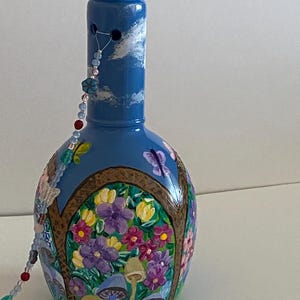 May include: A blue glass bottle with a painted scene of a garden with flowers, mushrooms, and butterflies. The bottle has a beaded garland hanging from the top.