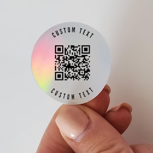 May include: A white iridescent sticker with a black and white QR code and the text "CUSTOM TEXT" printed around the code. The sticker is being held by a hand.