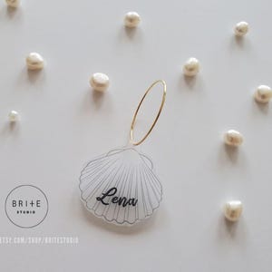May include: A clear acrylic seashell place card with a gold ring and the name "Lena" written in black. The place card is surrounded by white pearls.