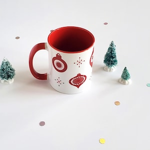 Könnte beinhalten: Eine weiße Keramiktasse mit rotem Innenraum und Henkel, verziert mit roten Weihnachtsschmuck-Designs. Kleine grüne Weihnachtsbäume und bunte Konfetti umgeben die Tasse und schaffen eine festliche Urlaubsszene.