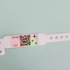 以下が含まれることがあります： QRコードと「CUSTOM」と「TEXT」の文字が印刷された白いリストバンド。 リストバンドには調整用の穴がいくつかあり、カラフルなグラデーションデザインです。 背景は明るいティール色です。