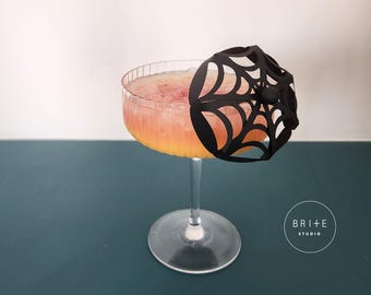 May include: A cocktail in a clear glass with a stem, filled with a pink and orange drink and ice. A black spiderweb-shaped decoration sits on the rim. The drink is on a teal surface.