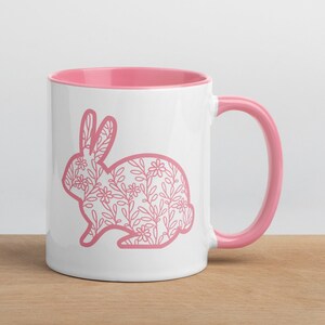 Könnte beinhalten: Eine weiße Keramiktasse mit rosafarbenem Rand und Henkel. Die Tasse zeigt eine rosafarbene Umrandung eines Kaninchens mit einem Blumenmuster.