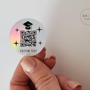 May include: A round, holographic sticker with a graduation cap icon, a QR code, and the words "CUSTOM TEXT." Four starbursts surround the design. The sticker has a rainbow gradient effect and is held by a person.