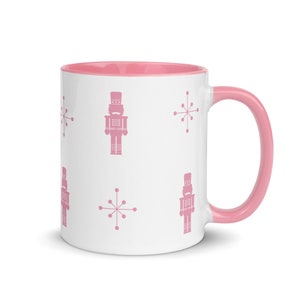 Könnte beinhalten: Weiße Keramiktasse mit rosa Innenraum und Griff. Die Tasse hat ein sich wiederholendes Muster aus rosa Nussknackern und Sternenmustern. Ein festlicher und dekorativer Artikel für Getränke.