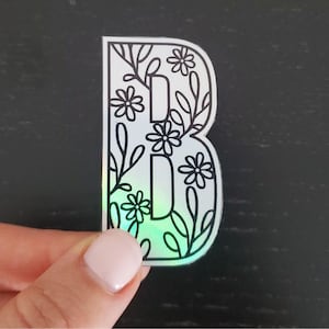 May include: A holographic sticker with a black outline of the letter "B" surrounded by flowers and leaves.