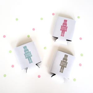 Puede incluir: Tres enfriadores de latas blancos con un diseño de cascanueces en rosa, gris y verde azulado. El fondo es blanco con lunares rosas y verdes. Los enfriadores están diseñados para mantener las bebidas frías.