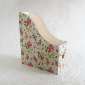 Puede incluir: Un porta revistas de madera con un patrón floral en tonos rosas y verdes. El porta revistas tiene un frente curvo y un agujero en el lateral.