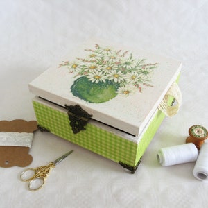 Può includere: Una scatola di legno a scacchi verde e bianca con un motivo floreale sul coperchio. Il coperchio presenta un bouquet di margherite bianche in un vaso verde. La scatola ha una chiusura in ottone e un manico a nastro bianco.