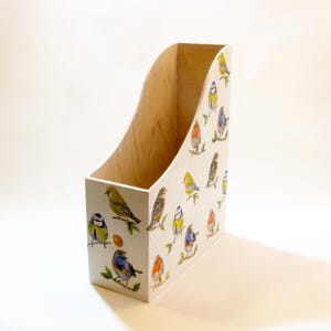 Op de afbeelding: Een houten tijdschriftenhouder met een gebogen bovenkant en een licht houten interieur. De witte buitenkant is versierd met kleurrijke illustraties van vogels. De vogels, in geel, blauw, oranje en bruin, zitten op takken.