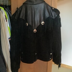 Puede incluir: Chaqueta de ante negra con flecos y botones decorativos plateados. La chaqueta presenta un canesú de cuero y flecos a lo largo de las mangas y la espalda. Una prenda de vestir elegante.