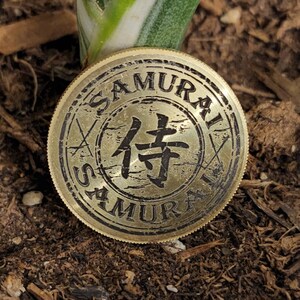 Può includere: Una moneta di metallo color oro con la parola "SAMURAI" incisa sul bordo. Il centro della moneta presenta un carattere giapponese in nero, circondato da uno sfondo testurizzato. Il carattere è "侍", che significa "samurai".