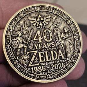 May include: A bronze-colored commemorative coin celebrating 40 years of The Legend of Zelda. The coin features detailed relief artwork of characters, a castle, and the Triforce symbol. The text "40 YEARS THE LEGEND OF ZELDA 1986-2026" is inscribed on the coin.