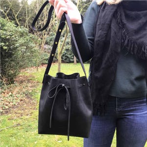 May include: A black bucket bag with a drawstring closure and a long adjustable shoulder strap featuring gold-toned metal rivets. The bag has a smooth matte finish and is constructed with a single main compartment and a cinched top opening.