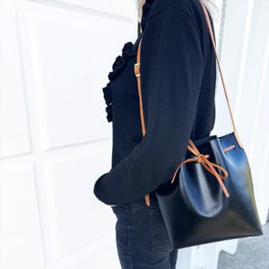 May include: A black bucket bag with a smooth finish, featuring a tan leather shoulder strap and a matching tan leather drawstring closure tied in a bow at the front. The bag has a structured base and is worn over the shoulder against a black top.