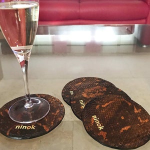 Könnte beinhalten: Vier braune Lederuntersetzer mit Schlangenhautmuster und goldener Beschriftung "ninok".