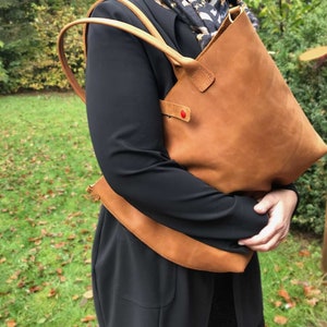 May include: A brown leather tote bag with a red heart detail. The bag has a long strap and is being held by a person.