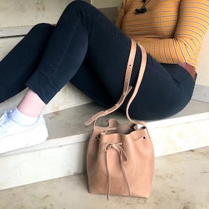 Può includere: Una borsa a secchiello in pelle marrone chiaro con chiusura a coulisse e una lunga tracolla. La borsa ha la parola "ninok" impressa sul davanti.