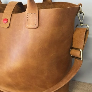 May include: A brown leather tote bag with a red button and a long strap. The bag has a top handle and a detachable shoulder strap.