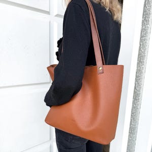 May include: A large, structured tote bag in a warm cognac brown colour with a pebbled texture. The bag features two long, flat shoulder straps attached with small, circular brass-toned metal rivets at the top corners of the main body.