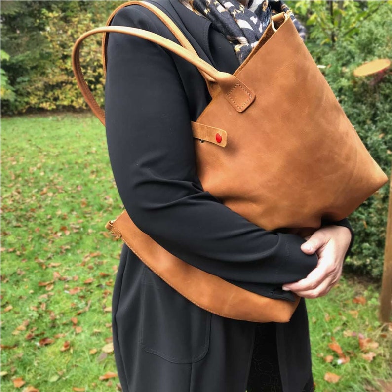 May include: A brown leather tote bag with a red heart detail. The bag has a long strap and is being held by a person.