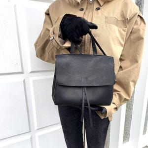 May include: A black leather backpack with a flap closure and two vertical straps. The bag has a top handle. The person holding the bag is wearing black gloves and a tan jacket.