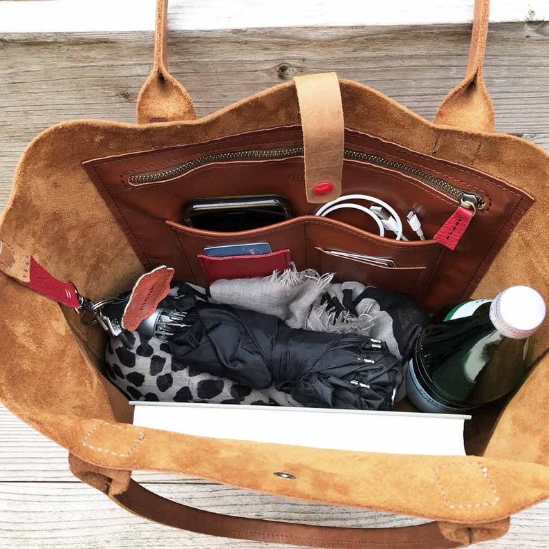 May include: A brown leather tote bag with multiple pockets and compartments. The bag is open and filled with items such as a phone, wallet, keys, a scarf, an umbrella, and a water bottle.