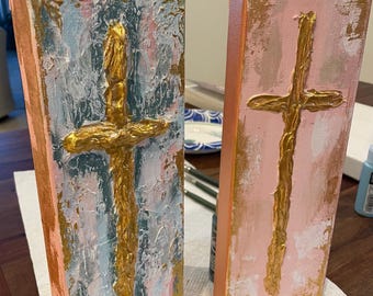 May include: Two abstract paintings of gold crosses on canvas. The cross on the left is on a blue and pink background. The cross on the right is on a pink background.