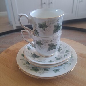 Op de afbeelding: Een witte theekop en schotelset met een groen klimopbladontwerp en gouden rand. De set bevat twee gestapelde kopjes en twee schotels. De set staat op een houten oppervlak.