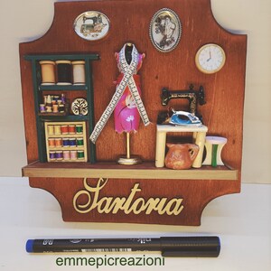 Op de afbeelding: Een houten bord met een miniatuur scène van een kleermaker. Het bord heeft een kleine naaimachine, spoelen garen, een paspop met een roze jurk en een bord met de tekst "Sartoria".