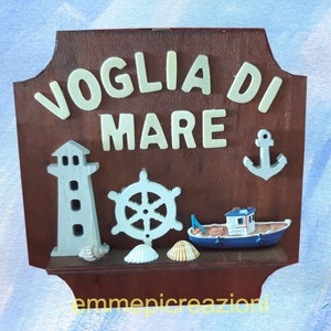 May include: A wooden sign with the words "VOGLIA DI MARE" in white letters. The sign is decorated with a lighthouse, a ship's wheel, a small boat, an anchor, and seashells. The text "emmepicreazioni" is at the bottom of the sign.
