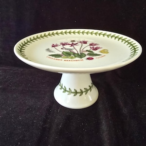 May include: A white ceramic cake stand featuring a botanical illustration of pink Cyclamen Repandum flowers, green leaves, and a small ladybug. The rim and base are decorated with a green leaf vine pattern, and the text Cyclamen Repandum is printed.