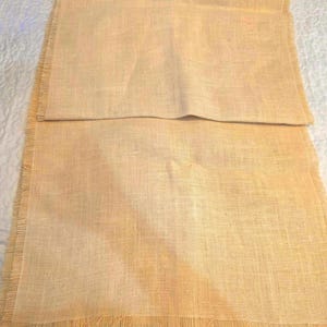May include: A rectangular table runner made of light tan, woven fabric with a visible coarse texture. The runner features frayed, unfinished edges along the sides and ends, creating a decorative fringe effect. A small white tag is attached at one corner.