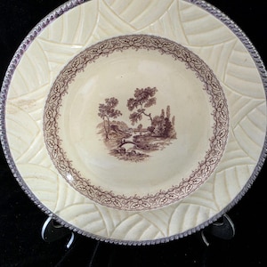 May include: A cream-coloured ceramic plate featuring a brown transferware landscape scene in the centre, surrounded by a decorative leaf border and a textured, basket-weave pattern on the rim with a silver-toned metallic edge trim.