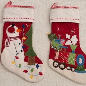 Puede incluir: Dos calcetines navideños rojos con puños acolchados blancos y ribete de cuadros rojos. Uno presenta un muñeco de nieve aplicado con bufanda y luces. El otro muestra un tren rojo y verde que transporta regalos envueltos en su vagón.