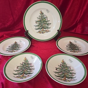Puede incluir: Un juego de cinco platos de cerámica blanca con borde verde y una ilustración central de un árbol de Navidad decorado con regalos debajo. La colección incluye un plato grande y cuatro cuencos más pequeños, todos con el mismo motivo de árbol.