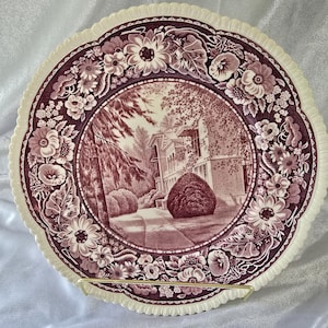 May include: A round ceramic plate with a scalloped edge featuring a deep plum-colored transferware pattern. The center depicts a detailed landscape with a house and trees, while the border is decorated with a dense arrangement of various flowers.