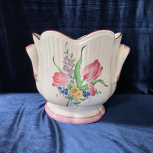 Puede incluir: Una maceta de cerámica color crema con borde y base festoneados, que presenta un motivo floral pintado a mano de tulipanes rosas, flores amarillas y hojas verdes. El recipiente tiene dos asas laterales con detalles en rosa y cuerpo acanalado.