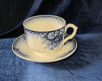 May include: A cream-colored ceramic teacup and matching saucer featuring a dark blue floral pattern. The rim of the cup and the edge of the saucer are decorated with a repeating arched border design, while the side displays detailed botanical illustrations.