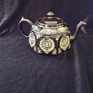May include: A dark brown hexagonal teapot featuring a glossy finish and intricate gold-colored filigree patterns. The design includes circular laurel wreath motifs on each side, a matching lid with a small knob, a curved handle, and a spout.