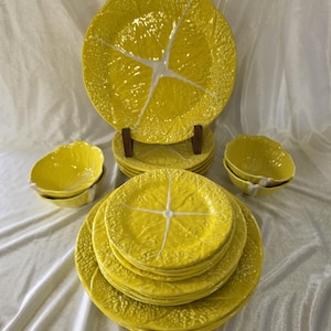 May include: A collection of bright yellow ceramic dinnerware featuring a textured cabbage leaf pattern with white vein details. The set includes a large serving plate on a stand, several smaller plates stacked in the center, and two small bowls.