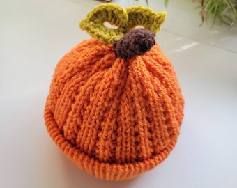 May include: An orange knit hat shaped like a pumpkin, with a brown stem and green leaves. The hat is made of a textured yarn and is likely handmade. The hat is a seasonal item, perfect for fall or Halloween.