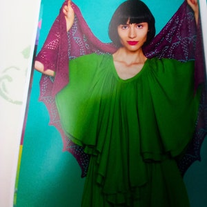 May include: A woman wearing a green dress with a purple and black lace shawl. The shawl is draped over her shoulders and arms. The woman has dark hair and is looking at the camera.