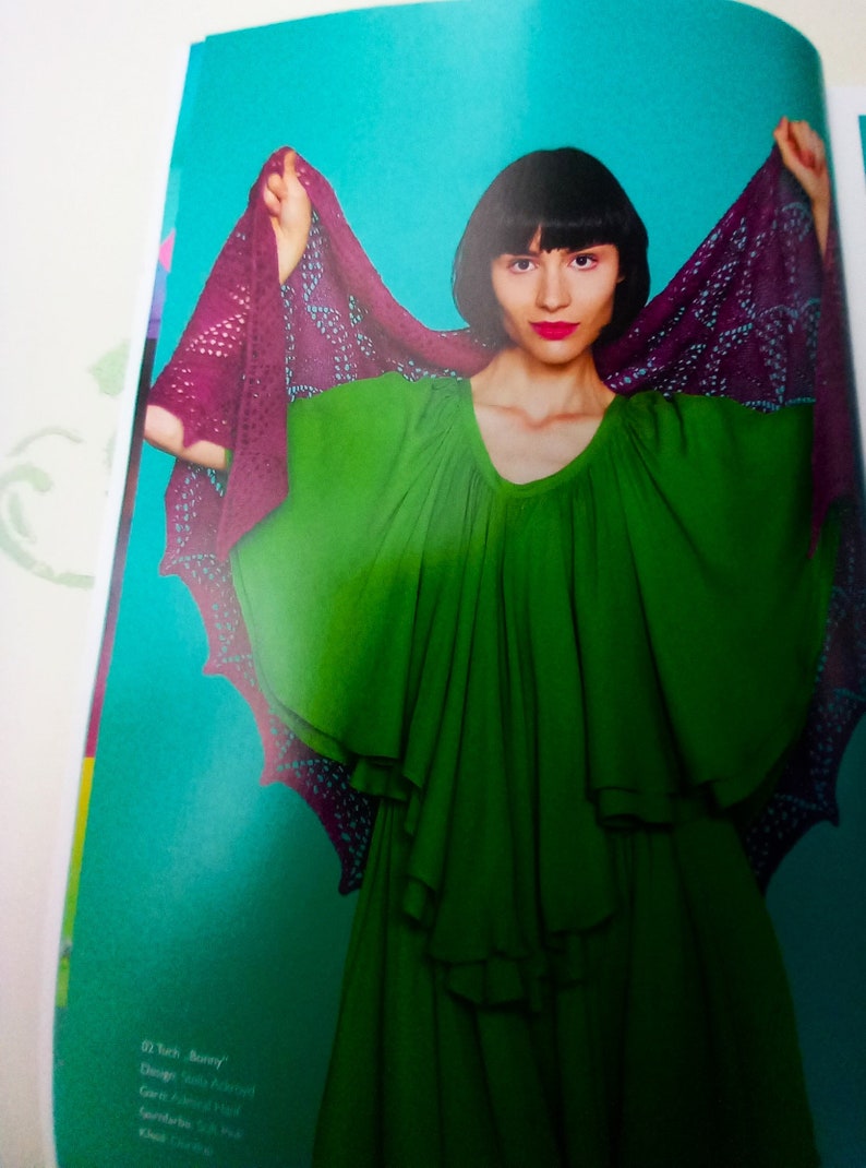 May include: A woman wearing a green dress with a purple and black lace shawl. The shawl is draped over her shoulders and arms. The woman has dark hair and is looking at the camera.