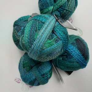 Pode incluir: Uma coleção de cinco novelos de lã redondos em vários tons de verde-azulado, verde-esmeralda e azul-escuro. Cada novelo apresenta um padrão de cor matizado e multicolorido com uma textura torcida. Várias etiquetas de papel preto estão presas.
