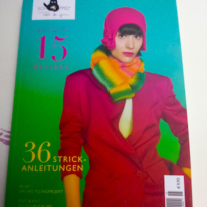 May include: A magazine cover featuring a woman wearing a pink hat and a colorful scarf. The cover text reads "15 MIRAKEL" and "36 STRICK-ANLEITUNGEN".