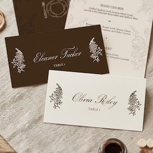 Puede incluir: Un tarjetón de mesa marrón oscuro con el nombre Eleanor Tucker y un diseño floral blanco, junto a un tarjetón color crema con el nombre Olivia Reiley y un diseño floral marrón, ambos con el texto TABLE 1 y un menú al fondo.