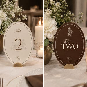 May include: Two oval table number signs standing on gold semi-circular bases. The left sign is cream with the text Table 2 and R&G. The right sign is dark brown with the text Table TWO and R&G, featuring a small floral line drawing above the text.