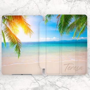May include: An open tablet case featuring a tropical beach scene. The design includes a sunny sky, turquoise water, white sand, and green palm fronds. The name "Teresa" is printed in a cursive font on the lower right side of the case.